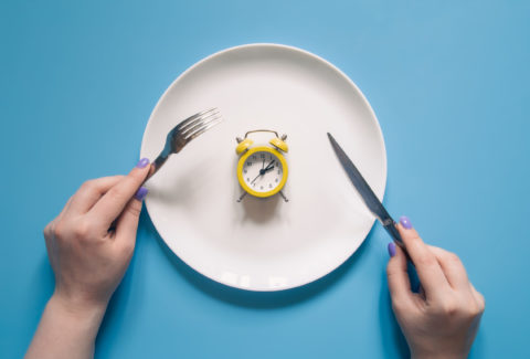 Hands holding knife and fork above alarm clock on a plate on blue background.