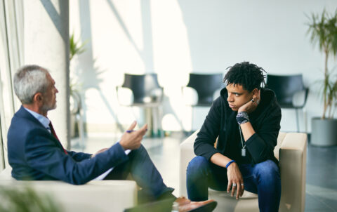 Black teenage boy communicating with mature psychotherapist during session at counselling center.