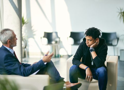 Black teenage boy communicating with mature psychotherapist during session at counselling center.