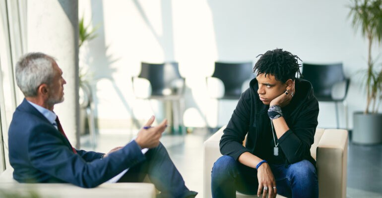 Black teenage boy communicating with mature psychotherapist during session at counselling center.